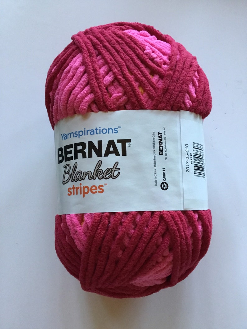 May include: A ball of pink and red yarn with a white label that reads "Yarnspirations&trade; BERNAT&reg; Blanket stripes&trade;". The label also includes the text "Made in China/Fabriqu&eacute; en Chine/Hecho en China", "CA00111", "BERNAT", "PO Box 40, Listowel, ON N4W 3J0", and "2017-05-010".