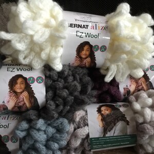 Puede incluir: Cinco ovillos de hilo Bernat Alize EZ Wool en varios colores, incluyendo blanco, gris, morado y azul. El hilo es de peso grueso y tiene una textura peluda. Cada ovillo tiene una etiqueta con el logotipo de Bernat Alize EZ Wool e instrucciones para usar el hilo.