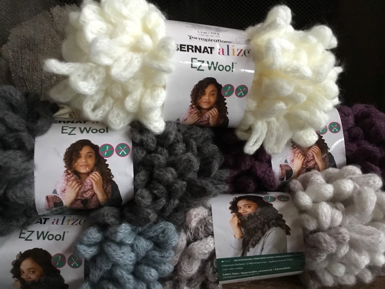 Puede incluir: Cinco ovillos de hilo Bernat Alize EZ Wool en varios colores, incluyendo blanco, gris, morado y azul. El hilo es de peso grueso y tiene una textura peluda. Cada ovillo tiene una etiqueta con el logotipo de Bernat Alize EZ Wool e instrucciones para usar el hilo.