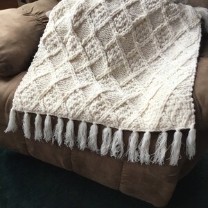 May include: A white knitted throw blanket with a diamond pattern and fringe trim. The blanket is draped over a brown armchair.
