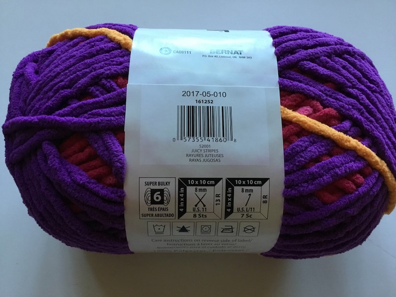 May include: A skein of Bernat Super Bulky yarn in the color Juicy Stripes. The yarn is a mix of purple, pink, and yellow. The label shows the yarn weight, needle size, and care instructions.