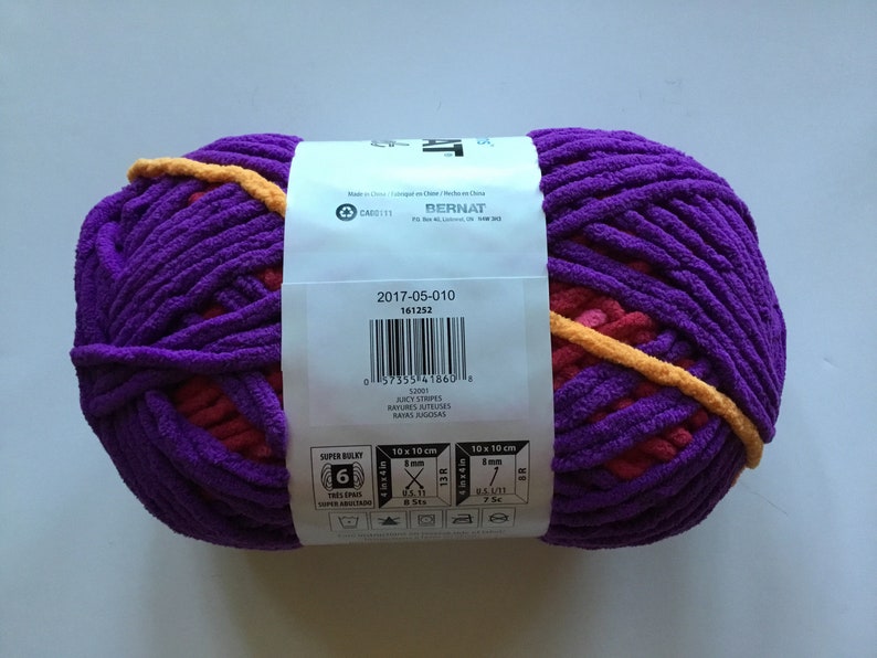 May include: A skein of Bernat Super Bulky yarn in the color Juicy Stripes. The yarn is a mix of purple, pink, and orange. The label includes the yarn weight, recommended needle size, and care instructions.