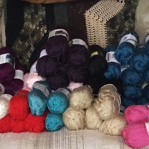 May include: A collection of colorful yarn skeins in various shades of green, red, blue, purple, and pink. The yarn is arranged in rows on a light brown surface.