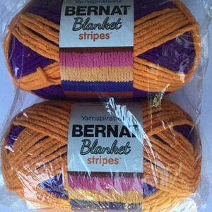 May include: Two skeins of Bernat Blanket Stripes yarn in orange, purple, pink, and yellow. The yarn is packaged in clear plastic.