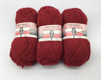 May include: Three balls of red heart Highlander knitting yarn. The yarn is a deep red color with a subtle fleck of blue. The label reads "Red Heart Highlander Knitting Worsted Tricot Mode."