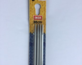 May include: A package of silver knitting needles. The label reads "3" and "INOX 30cm". The packaging has a yellow and blue knitted pattern design. The needles are made of metal.