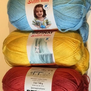 May include: A stack of six balls of Bernat Softee Baby yarn in various colours, including green, blue, yellow, red, grey, and multi-coloured. Each ball is labelled with the brand name, yarn type, and weight. The yarn is 100% acrylic and comes in 5 oz (140 g) balls.