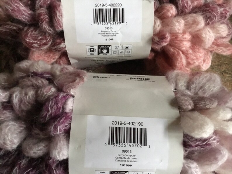 Puede incluir: Dos ovillos de hilo Bernat, uno en una combinaci&oacute;n de colores rosa y blanco llamada "Burgundy Peony" y otro en una combinaci&oacute;n de colores morado y blanco llamada "Berry Compote". Ambos ovillos tienen un aspecto peludo y texturizado.