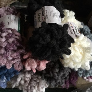 Puede incluir: Primer plano de varios ovillos de hilo Bernat EZ Wool en varios colores, incluyendo morado, negro, blanco, gris y azul. El hilo tiene una textura gruesa y peluda.