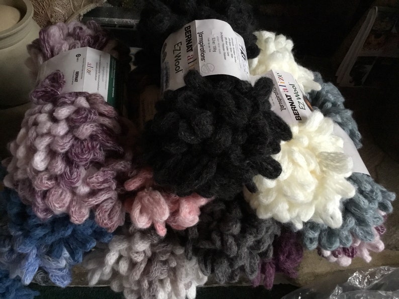 Puede incluir: Primer plano de varios ovillos de hilo Bernat EZ Wool en varios colores, incluyendo morado, negro, blanco, gris y azul. El hilo tiene una textura gruesa y peluda.