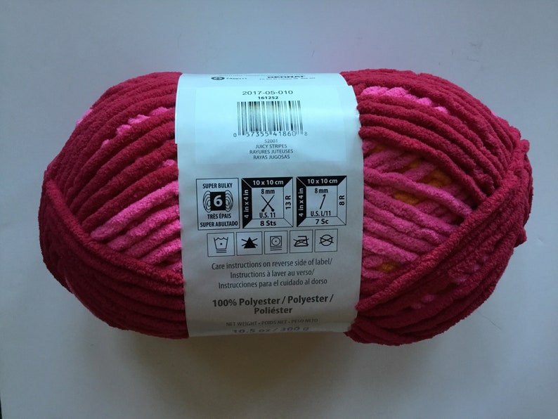 May include: A ball of pink and orange super bulky yarn with a label that says "Juicy Stripes" and "100% Polyester". The label also includes care instructions and knitting gauge information.