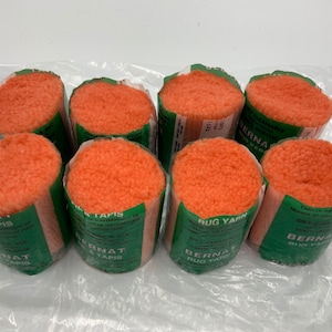 May include: Eight spools of orange Bernat Rug Yarn. The spools are wrapped in clear plastic and have green labels with the text "Bernat Rug Yarn" and "Tapis" in multiple languages.