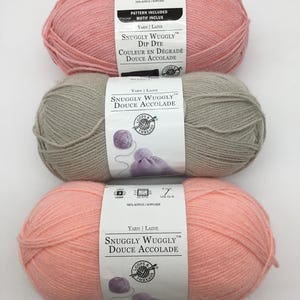 May include: Three balls of yarn in pink, grey, and peach colours. The yarn is labelled "Snuggly Wuggly" and "Douce Accolade". The yarn is 100% acrylic.