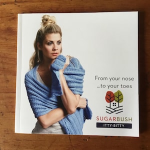 May include: A woman wearing a light blue knitted shawl with the text "From your nose...to your toes" and the logo "Sugarbush Itty-Bitty"