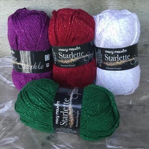 May include: Four skeins of Mary Maxim Starlette Sparkle yarn in purple, red, white, and green. Each skein is labelled with the brand name and yarn type. The yarn has a sparkly, metallic thread woven throughout.