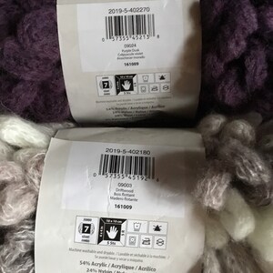 Puede incluir: Dos ovillos de hilo, uno morado y otro marr&oacute;n y blanco, con etiquetas que muestran el nombre, el color y el contenido de la fibra del hilo. Las etiquetas tambi&eacute;n incluyen instrucciones de cuidado y el peso del hilo. El hilo se llama "Yarnspirations" y es un hilo de peso jumbo.