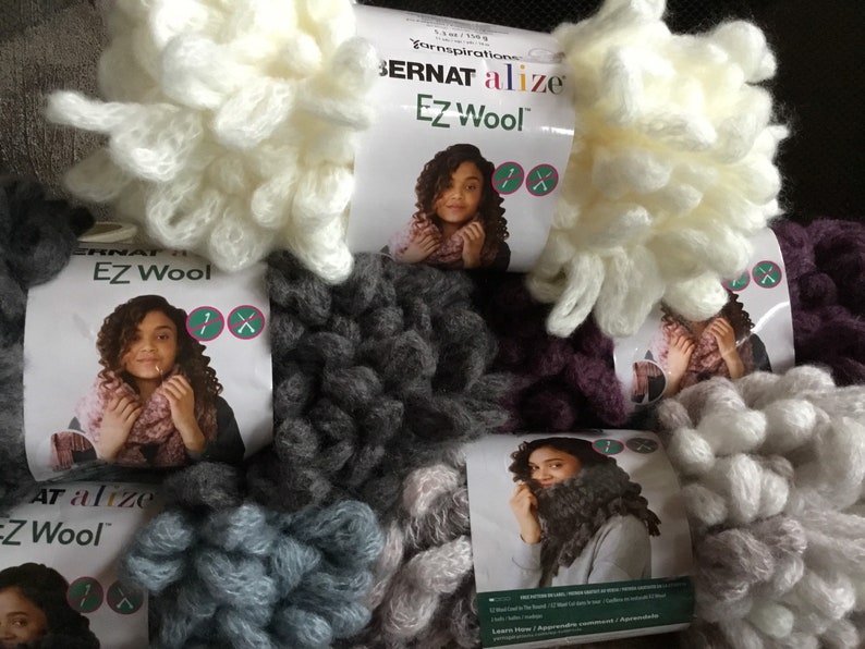 Puede incluir: Primer plano de varios ovillos de hilo Bernat Alize EZ Wool en varios colores, incluyendo blanco, gris, morado y azul. El hilo es de peso grueso y tiene una textura peluda. Las etiquetas de hilo muestran a una mujer usando una bufanda hecha con el hilo.