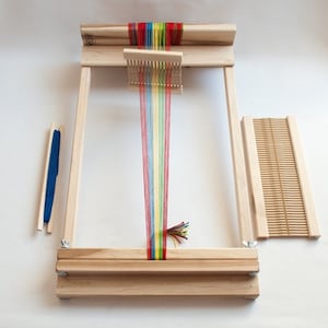 May include: A wooden loom with a rainbow colored yarn threaded through it. The loom has a wooden frame and a shuttle for weaving. There is also a separate wooden comb for finishing the woven fabric.