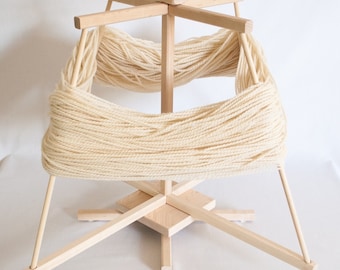 Puede incluir: Un enhebrador de lana de madera con una bola de lana blanca enrollada alrededor. El enhebrador tiene cuatro brazos y una base giratoria.