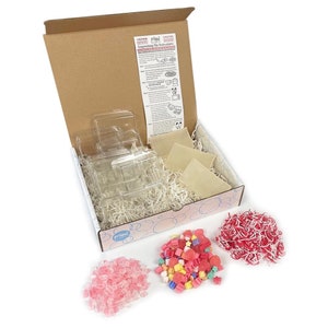 May include: A box filled with supplies for making soap. The box contains clear plastic molds, white soap base, pink and white shredded paper, and colorful soap pieces.
