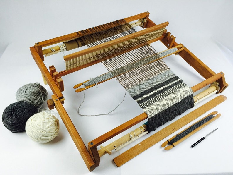 May include: A wooden loom with a partially woven tapestry in shades of grey and white. Three balls of yarn in grey and white are in the foreground.