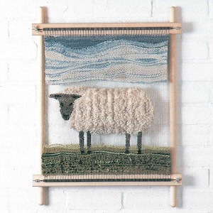 May include: A woven wall hanging featuring a sheep design. The sheep has a fluffy, cream-colored body and a dark head and legs. The background depicts a blue and white sky above a green field, all within a wooden frame.