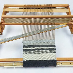May include: A wooden loom with a grey and white striped woven fabric on it. The loom has a wooden frame and a wooden shuttle.