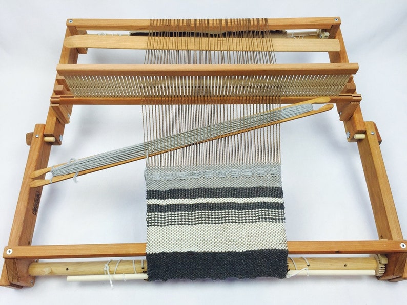 May include: A wooden loom with a grey and white striped woven fabric on it. The loom has a wooden frame and a wooden shuttle.