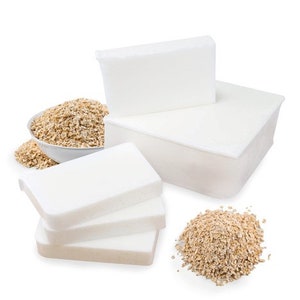 May include: Three white rectangular bars of soap, two stacked on top of each other, and one lying flat on the table. A white rectangular container with a white lid is behind the soap bars. Two piles of oatmeal are on the table, one in a white bowl and the other loose.