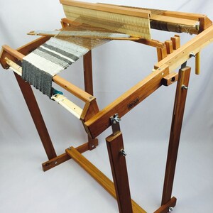 May include: A wooden loom with a grey and white striped fabric woven on it. The loom is on a stand with a wooden frame and metal hardware.