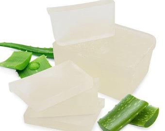 May include: Four clear, translucent bars of aloe vera soap stacked on top of each other. The soap is surrounded by green aloe vera leaves.