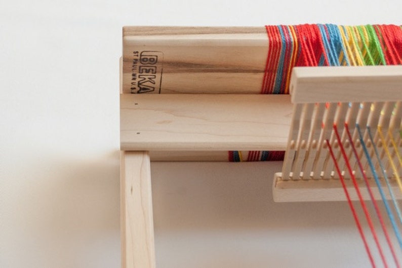May include: A wooden loom with colorful threads strung across it. The loom is made by BEKA ST. PAUL, MN, U.S.A.