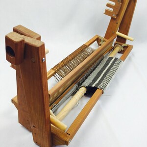 May include: A wooden loom with a partially woven rug on it. The loom is made of natural wood and has a simple design. The rug is a striped pattern in grey and white.