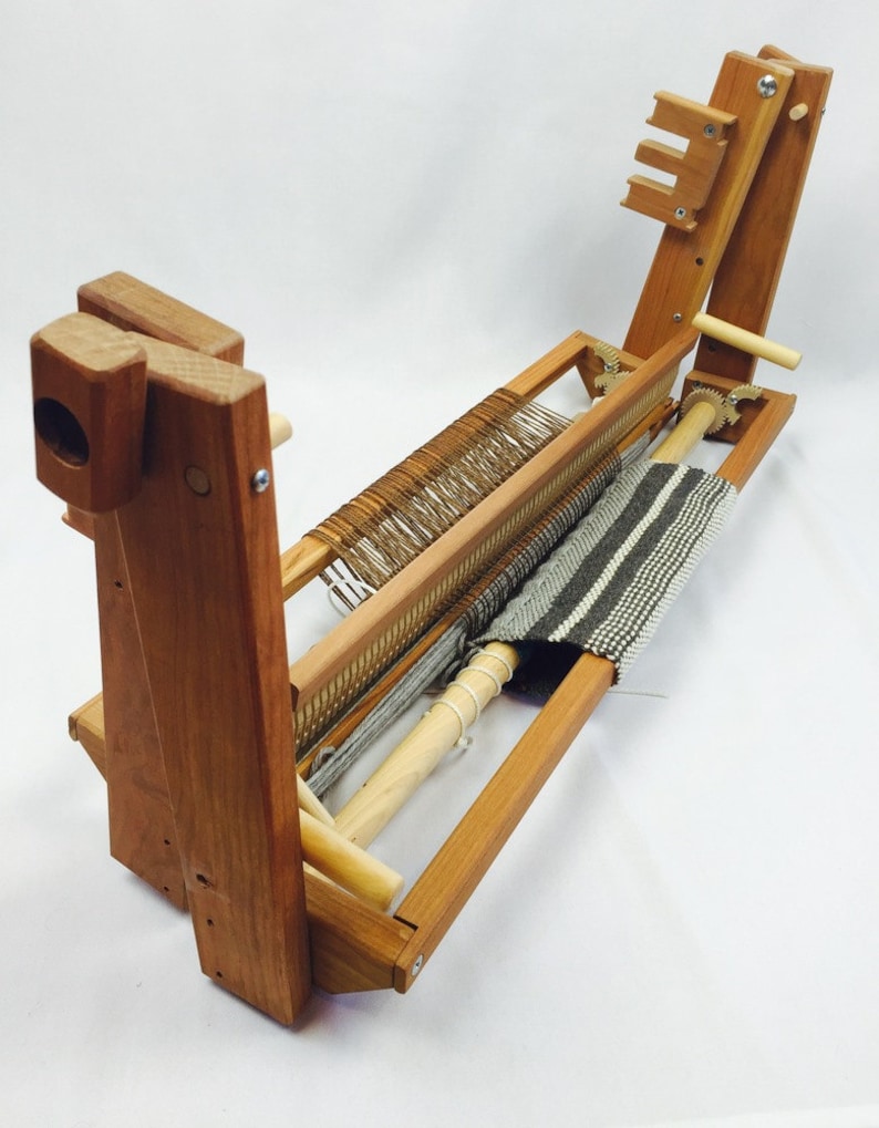 May include: A wooden loom with a partially woven rug on it. The loom is made of natural wood and has a simple design. The rug is a striped pattern in grey and white.