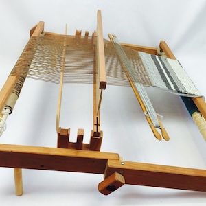 May include: A wooden loom with a partially woven fabric in a striped pattern. The loom has a wooden frame and a wooden shuttle. The loom is set up on a wooden stand.