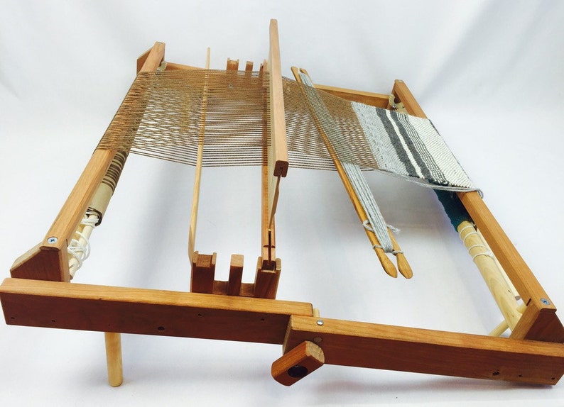 May include: A wooden loom with a partially woven fabric in a striped pattern. The loom has a wooden frame and a wooden shuttle. The loom is set up on a wooden stand.