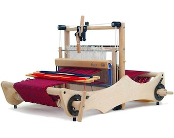 Puede incluir: Un telar de madera con una tela tejida roja, azul y blanca. El telar tiene un marco de madera y una manivela de metal. El telar está etiquetado como "Erica".