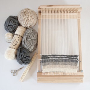 May include: A wooden loom with a woven tapestry in shades of gray and beige.  The loom is surrounded by balls of yarn in various shades of gray and beige.  The yarn is labeled "The Woolery Co."