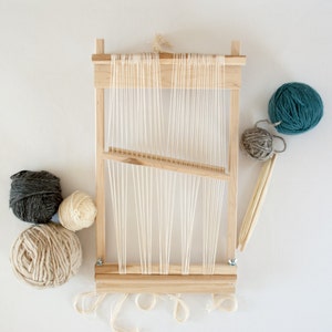 May include: A wooden loom with white yarn strung across it. There are four balls of yarn in different colors: gray, beige, teal, and gray.