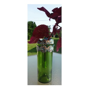 May include: A green glass bottle vase with a decorative mosaic trim around the top. The vase is filled with a single stem of purple foliage.