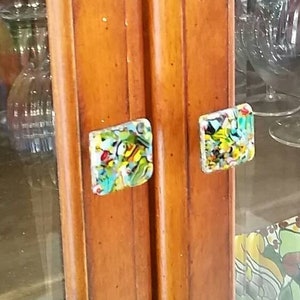 Könnte beinhalten: Ein Holzschränkchen mit Glastüren und Regalen. Der Schrank hat zwei bunte Glasknöpfe. Die Regale sind mit Glaswaren gefüllt, darunter eine Karaffe und Gläser. Auf dem untersten Regal steht eine bunte Keramikkaraffe.