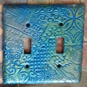 Op de afbeelding: Een blauwe en groene lichtschakelaar met een decoratief reliëfpatroon. Het patroon omvat swirls, sterren en geometrische patronen. De lichtschakelaar heeft twee schakelaars.