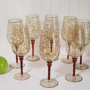 Pode incluir: Um conjunto de oito taças de vinho com taças âmbar e hastes vermelhas. As taças apresentam um delicado padrão de filigrana dourada. Uma maçã verde está posicionada à esquerda das taças.