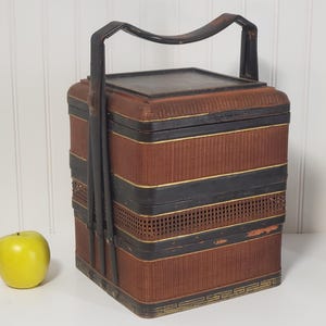 May include: A vintage brown and black woven basket with a handle. The basket has a rectangular shape with black bands and gold accents. A green apple sits next to the basket.
