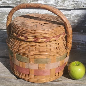 May include: A round, lidded wicker basket with a woven handle. The basket features bands of pink and green woven into the natural tan color. A green apple sits next to the basket.