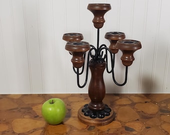 May include: A five-arm candelabra featuring a central wooden pillar and base with black metal curved arms supporting five dark brown wooden candle cups. The piece is positioned next to a single green apple for scale, showcasing its height and structure.