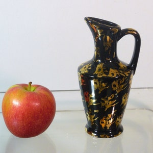 Puede incluir: Una pequeña jarra de cerámica negra con detalles en pan de oro. La jarra tiene una boca ancha y un asa. Está sobre una superficie de vidrio junto a una manzana roja.