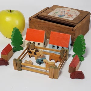 May include: A wooden toy farm set with a yellow apple. The set includes miniature houses with orange roofs, green trees, a wooden fence, and various animal figures. A wooden box with a lid is also present.