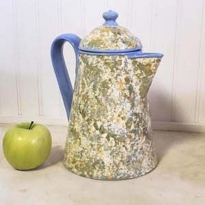 May include: A ceramic pitcher with a lid, featuring a mottled sponge-painted pattern in shades of sage green, yellow, and light blue. The handle, lid knob, and rim are finished in a solid light blue glaze. A single green apple sits beside it.
