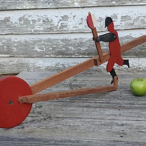 May include: A vintage wooden toy featuring a red wheel, a brown frame, and a red figure with a black hat. The figure appears to be pushing a pole with a red and white top. A green apple rests nearby.
